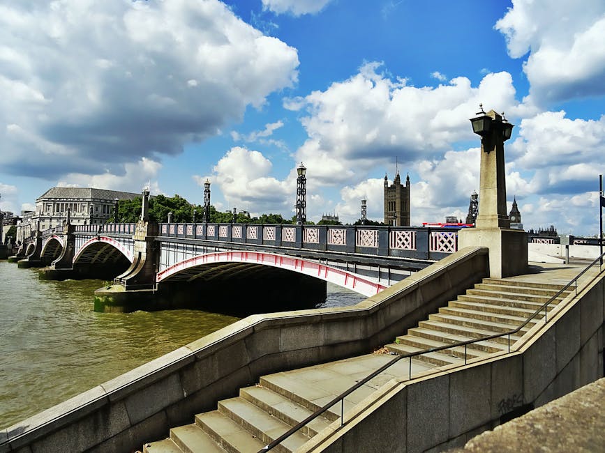 A stone staircase with metal handrails leading down to a riverside walkway in Lambeth, featuring clean, smooth stone steps and a well-maintained balustrade. The scene includes a view of Westminster Bridge with its red and white-painted ironwork arches spanning the river Thames, under a partly cloudy sky with sunlight illuminating the area. In the background, the historic architecture of the Palace of Westminster is visible, along with the iconic clock tower of Big Ben. The surface appears clean and dry, emphasizing the importance of regular surface cleaning and maintenance by professionals like Cleaners Lambeth, who provide expert domestic and commercial cleaning services in the area.