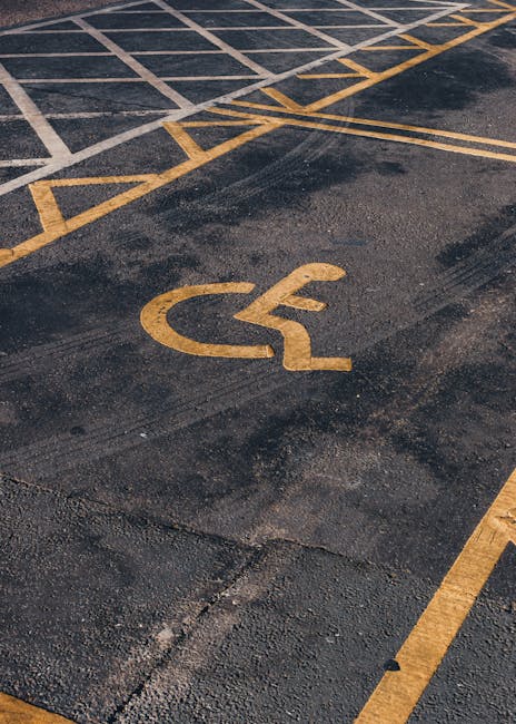 A close-up view of an outdoor parking area surface with a yellow wheelchair accessibility symbol painted on dark asphalt. The ground appears slightly worn but is generally clean, with visible white and yellow parking lines creating designated spaces and pathways. The surface shows some tire marks and minor cracks, typical of outdoor parking lots. The lighting is natural, indicating daylight, and the area is clear of debris. This image reflects the importance of proper surface maintenance and cleanliness in parking areas, which is relevant to the deep cleaning and sanitisation services provided by Cleaners Lambeth to ensure accessible and hygienic surroundings for residents and visitors.