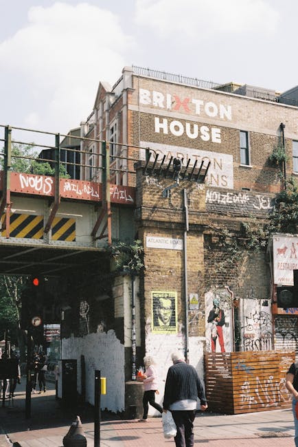 The image depicts an exterior view of the Brixton Market building, featuring a multi-story brick facade with large painted signage that reads 'BRIXTON HOUSE.' The building has a weathered, slightly aged appearance with visible graffiti and posters on the lower walls. A metal fire escape with a ladder extends from the rooftop to the awning, and the structure is adorned with greenery climbing the walls. In the foreground, pedestrians are walking beneath the market archway, including a person in a black jacket and others casually dressed, with some carrying shopping bags. The lighting is natural daylight, casting soft shadows and highlighting the textures of the brickwork and street environment. As part of professional cleaning services offered by Cleaners Lambeth, the area appears well-maintained with no visible dirt or debris, emphasizing the importance of surface cleaning and maintenance for such busy commercial locations.
