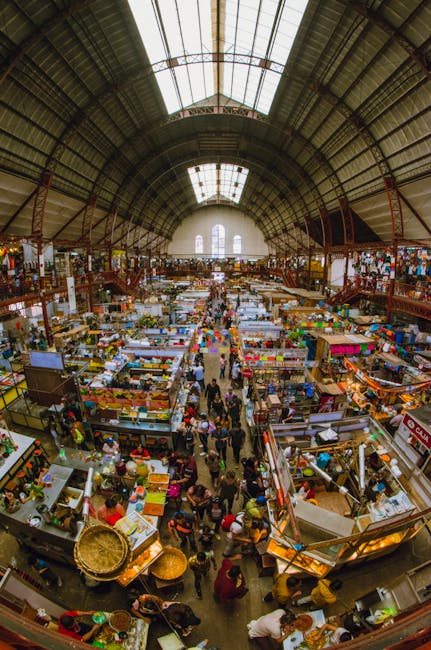 An indoor market with a large arched ceiling featuring skylights allowing natural light to illuminate the space. The market is filled with numerous vendor stalls arranged in rows, each displaying various goods such as textiles, produce, and household items. Shoppers browse and walk through the central aisle, which is busy with visitors. The surfaces, including wooden countertops and ceramic tiles, appear clean and well-maintained, reflecting professional deep cleaning. Visible cleaning tools like cloths and sprays suggest ongoing maintenance, contributing to a hygienic environment. The lighting highlights the cleanliness and orderliness of the market, emphasizing its vibrant yet orderly condition, consistent with commercial cleaning standards. This image is related to professional cleaning services for market spaces, as promoted by Cleaners Lambeth, who offer surface cleaning, deep cleaning, and sanitisation for commercial environments like markets.
