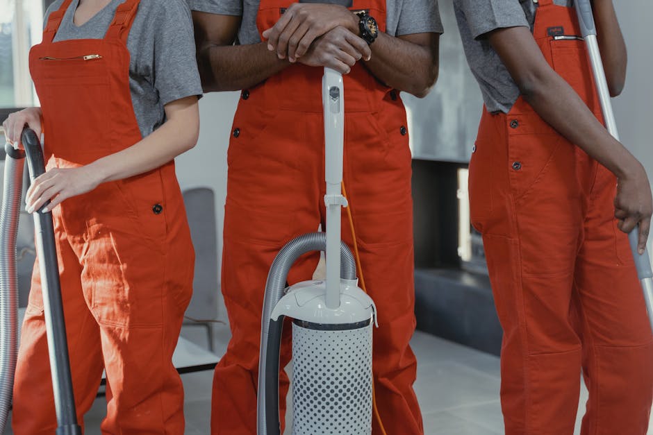 Two professional cleaners from Cleaners Lambeth stand back-to-back against a plain white wall in a well-lit room. The cleaner on the left, an Asian woman, holds a vacuum cleaner with a black hose and 