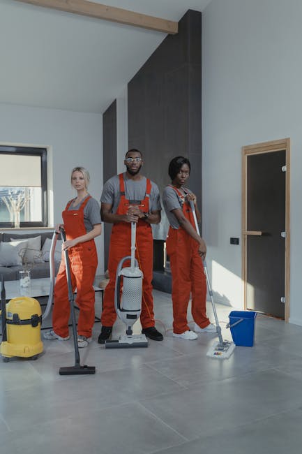 Three professional cleaners from Cleaners Lambeth are standing in a modern living room with light grey walls and large windows allowing natural light to illuminate the space. They are dressed in grey t-shirts and bright orange overalls, each holding cleaning tools: the first with a vacuum cleaner, the second with a spray bottle and cloth, and the third with a mop and bucket. The room features a dark wood accent wall, a grey tiled floor, and minimal furniture including a grey sofa and a side table with a bottle of water. Cleaning equipment such as a yellow vacuum machine and a blue mop bucket are visible on the floor, indicating they are preparing for or in the process of deep cleaning or surface sanitisation. Cleaners Lambeth specializes in domestic cleaning and surface maintenance, ensuring environments are hygienic and well-maintained.