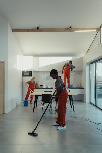 A modern, spacious kitchen with light grey tiled flooring and white walls, illuminated by natural light through large glass sliding doors. Two cleaning professionals from Cleaners Lambeth, wearing red aprons over casual clothes, are actively engaged in deep cleaning tasks. One is vacuuming the light-colored wooden dining table while another stands on a step ladder, polishing the high cabinets near the ceiling. A third worker in the background is mopping the floor, with a blue bucket nearby. The scene highlights thorough surface cleaning and maintenance of a contemporary domestic space, ensuring hygiene and shine across surfaces, and showcasing professional cleaning processes in a well-lit environment.