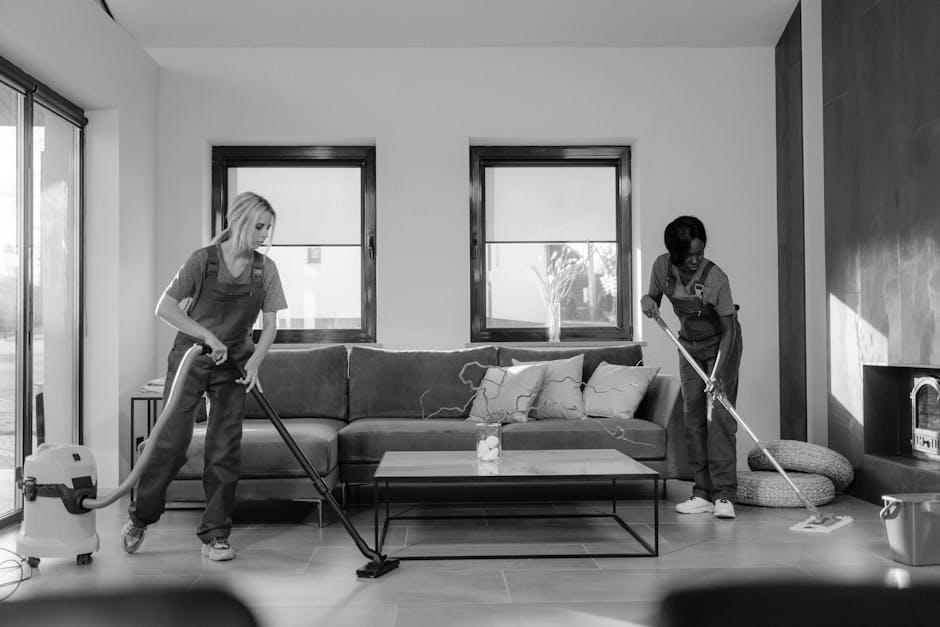 Three professional cleaners from Cleaners Lambeth are standing in a modern living room with light grey walls and large windows allowing natural light to illuminate the space. They are dressed in grey t-shirts and bright orange overalls, each holding cleaning tools: the first with a vacuum cleaner, the second with a spray bottle and cloth, and the third with a mop and bucket. The room features a dark wood accent wall, a grey tiled floor, and minimal furniture including a grey sofa and a side table with a bottle of water. Cleaning equipment such as a yellow vacuum machine and a blue mop bucket are visible on the floor, indicating they are preparing for or in the process of deep cleaning or surface sanitisation. Cleaners Lambeth specializes in domestic cleaning and surface maintenance, ensuring environments are hygienic and well-maintained.