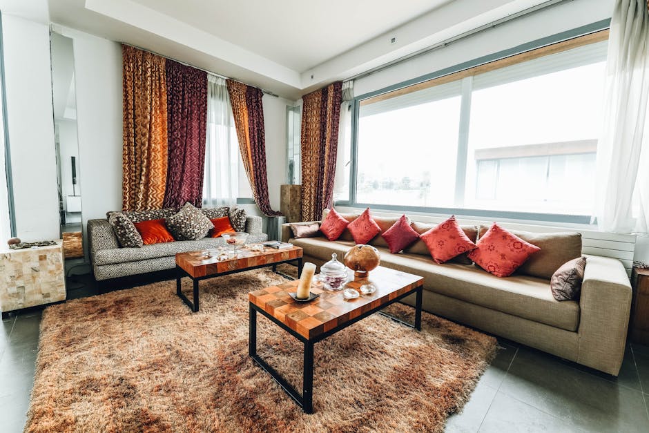 A spacious living room with a large beige sofa adorned with multiple red and patterned cushions, positioned along a wall of large windows with sheer white curtains and multicolored drapes. Two wooden coffee tables with black metal legs sit on a plush, textured beige area rug. The tables display decorative items such as candles, glass jars, and a wooden sphere. The light from the windows illuminates the room, highlighting the clean and tidy surface condition. The room features a mix of textured and smooth surfaces, including ceramic floor tiles and fabric upholstery, suggesting a focus on surface cleaning and maintenance by Cleaners Lambeth, the professional cleaning service. The overall appearance is well-maintained, emphasizing hygiene and deep cleaning in a domestic setting.