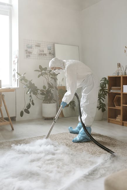A professional cleaner wearing a white protective suit, gloves, mask, and blue shoe covers is vacuuming a beige textured carpet in a bright, modern living room. The room features white walls, a large window allowing natural light, and potted plants adding greenery. There is a wooden sideboard on the right with decorative items, and a small desk with a stool near the window. Visible cleaning equipment includes a black vacuum hose. The scene emphasizes comprehensive carpet deep cleaning and sanitisation services provided by Cleaners Lambeth, promoting hygiene and maintenance of residential spaces in Lambeth SE11.