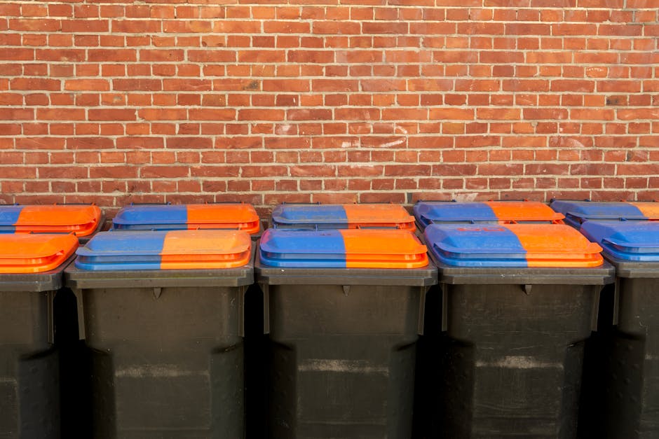 A man wearing a gray beanie, yellow safety vest, and black clothing is standing on a clean, tiled platform at a modern train station, next to a large hand truck loaded with black, orange, and white ga