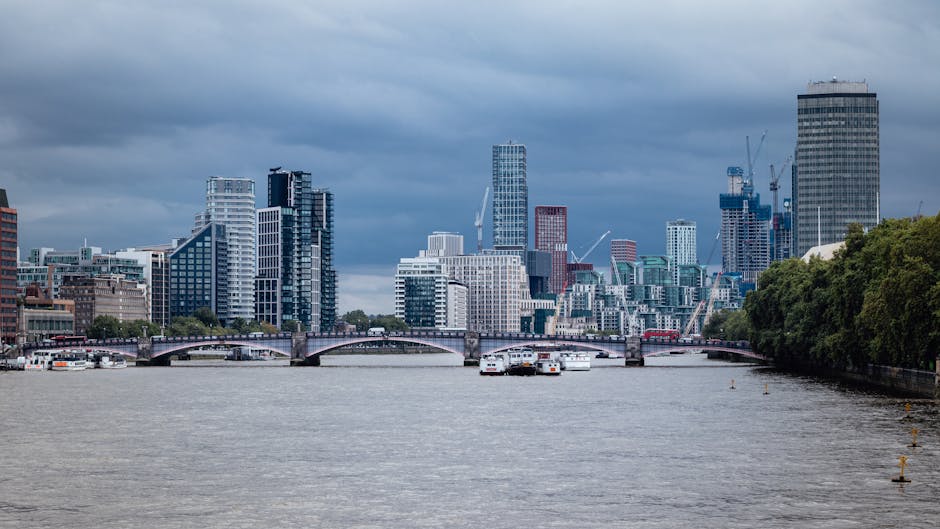 A panoramic view of the London skyline featuring modern high-rise buildings and skyscrapers, situated along the River Thames with a bridge spanning across the water. The river surface appears calm with some boats docked along the banks. The image has overcast weather with dark clouds overhead, demonstrating urban landscape and cityscape scenery typical of Lambeth, London. The scene emphasizes clean, well-maintained surfaces and reflective glass on the buildings, with lush greenery lining the riverside. Cleaners Lambeth specializes in surface cleaning and deep cleaning services aimed at maintaining hygiene and cleanliness in urban environments, ensuring appearances like this are preserved.