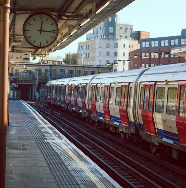 A modern train at an elevated urban train station platform, featuring a sleek stainless steel body with red and blue accents along the sides. The platform surface is made of concrete tiles, partially illuminated by overhead lighting, with tactile paving strips near the edge for safety. A large round clock shows the time, attached to the station ceiling, which also has visible wiring and lighting fixtures. In the background, multi-storey modern buildings with windows and balconies are visible under clear skies. This scene reflects the clean, well-maintained environment typical of urban surface cleaning and station hygiene management that companies like Cleaners Lambeth may support through their domestic and commercial sanitisation and deep cleaning services.