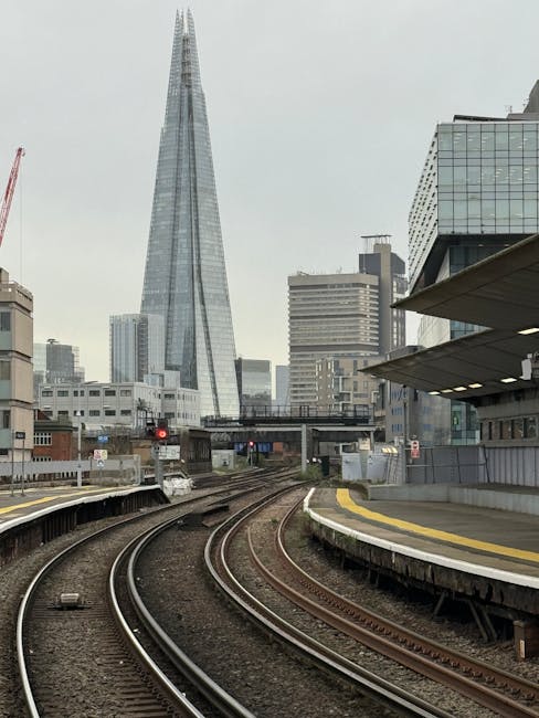 A view of train tracks on a curved section of railway with a modern high-rise building, known as The Shard, in the background. The tracks are clean and free of debris, with smooth steel rails and evenly spaced wooden sleepers. Adjacent to the tracks is a concrete platform with a yellow tactile strip along the edge, showing no signs of dust or clutter. To the right, a contemporary building with glass windows and awnings is visible. The scene is illuminated by natural daylight, although slightly overcast, emphasizing the cleanliness and well-maintained appearance of the railway area. This image reflects a focus on surface cleaning and maintenance, consistent with professional cleaning services like those provided by Cleaners Lambeth for residential or commercial properties near Waterloo Station, Lambeth.