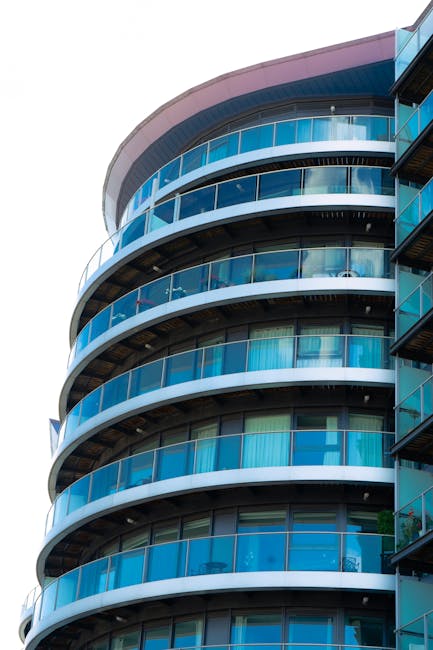 A modern multi-storey residential building featuring curved glass balconies with blue-tinted railings. The exterior surfaces include glass panels and concrete features, all appearing clean and well-maintained. The glass balcony doors reflect the surrounding environment, and natural light enhances the building's sleek design. The image showcases the building's architectural details and the pristine condition of its surfaces, indicative of professional deep cleaning and surface maintenance by Cleaners Lambeth as part of their domestic cleaning and sanitation services. The overall scene emphasizes hygiene, surface care, and building upkeep in a residential setting.
