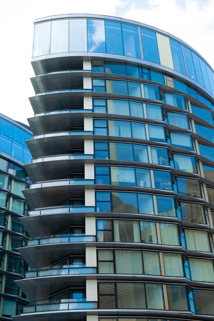 A close-up view of a modern high-rise building with curved glass balconies, featuring large glass windows and metallic railings. The building's exterior is composed of reflective blue-tinted glass panels, highlighting the sleek and contemporary architectural design. Bright daylight illuminates the facade, with reflections of the sky visible on the glass surfaces. This image emphasizes urban residential architecture typical of commercial and domestic spaces in Lambeth. For thorough surface cleaning, deep cleaning, and sanitisation of such buildings' exterior glass and balcony surfaces, professionals at Cleaners Lambeth are equipped to ensure maximum hygiene and shine on residential and commercial properties, aligning with the top tips for deep cleaning in the Vauxhall apartment context.